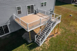 Aerial view of a wooden deck attached to a gray house with metal railings and stairs, surrounded by green lawn on a sunny day.