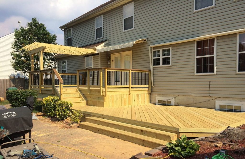 Newly constructed wooden deck with pergola attached to a two-story residential home with tan siding, featuring black metal railings and multiple levels.