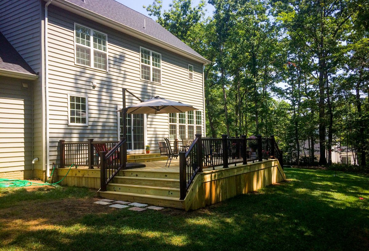 Suburban two-story house with cream siding and dark roof, featuring a wooden deck with black railings and umbrella, surrounded by mature trees and green lawn in bright daylight.